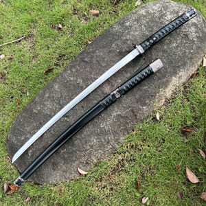 Op de afbeelding: Een Japanse katana-zwaard met een zilveren lemmet en zwarte handgreep en schede. Het zwaard ligt op een grote grijze rots, omringd door groen gras. De handgreep is gewikkeld in een getextureerd zwart materiaal.