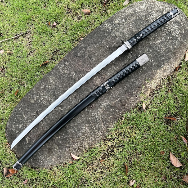 Op de afbeelding: Een Japanse katana-zwaard met een zilveren lemmet en zwarte handgreep en schede. Het zwaard ligt op een grote grijze rots, omringd door groen gras. De handgreep is gewikkeld in een getextureerd zwart materiaal.