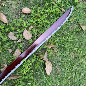 Peut inclure: Une lame d'&eacute;p&eacute;e courbe pr&eacute;sentant une finition rouge profond avec un motif g&eacute;om&eacute;trique sombre r&eacute;p&eacute;titif sur le plat. La lame poss&egrave;de un tranchant argent&eacute; m&eacute;tallique avec une ligne de hamon ondul&eacute;e s&eacute;parant la surface rouge de l'acier.