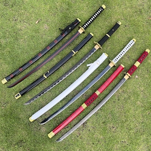 May include: Five different katana swords with black, white, and red scabbards. The swords have gold accents and are arranged on a green grassy surface.