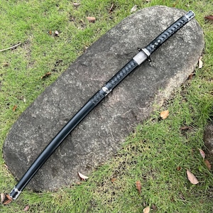 Op de afbeelding: Een zwart katana-zwaard met een getextureerde handgreep en een zilveren beschermer rust op een grote grijze rots. Het lemmet van het zwaard zit in een zwarte schede. De achtergrond heeft groen gras.
