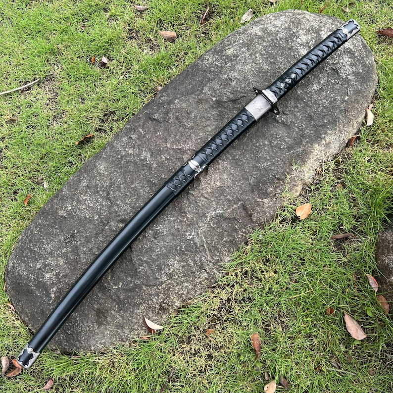 Op de afbeelding: Een zwart katana-zwaard met een getextureerde handgreep en een zilveren beschermer rust op een grote grijze rots. Het lemmet van het zwaard zit in een zwarte schede. De achtergrond heeft groen gras.
