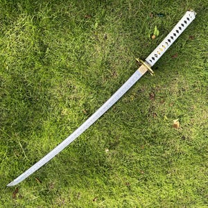 May include: A white katana sword with a black and gold patterned hilt. The sword is lying on a bed of green grass.
