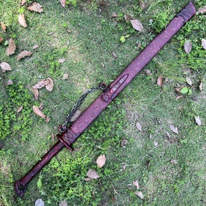 Peut inclure: Une &eacute;p&eacute;e longue avec un fourreau m&eacute;tallique rouge fonc&eacute; orn&eacute; de motifs en relief complexes et d'une petite pierre pr&eacute;cieuse rouge. La poign&eacute;e est &eacute;galement rouge fonc&eacute; avec des d&eacute;tails textur&eacute;s, et une cha&icirc;ne m&eacute;tallique argent&eacute;e est fix&eacute;e.
