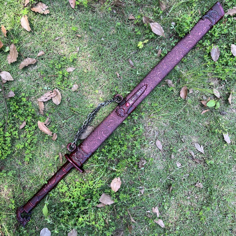 Peut inclure: Une &eacute;p&eacute;e longue avec un fourreau m&eacute;tallique rouge fonc&eacute; orn&eacute; de motifs en relief complexes et d'une petite pierre pr&eacute;cieuse rouge. La poign&eacute;e est &eacute;galement rouge fonc&eacute; avec des d&eacute;tails textur&eacute;s, et une cha&icirc;ne m&eacute;tallique argent&eacute;e est fix&eacute;e.