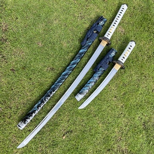 Op de afbeelding: Drie Japanse zwaarden met zwart-blauwe gepatroneerde handvatten en zilveren messen. De zwaarden liggen op een groene grasmat.