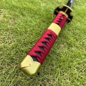 May include: Close-up of a red and black sword scabbard with a gold metal hilt. The scabbard is wrapped in red cord and has a black fabric underneath.