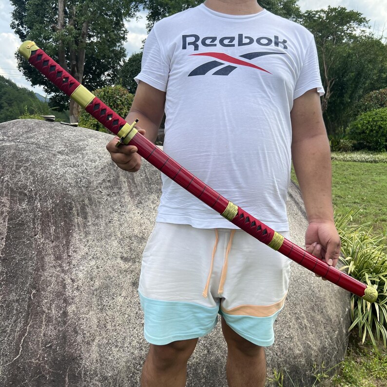 May include: A person holding a red and gold katana sword with a black handle. The sword has a decorative pattern on the blade.