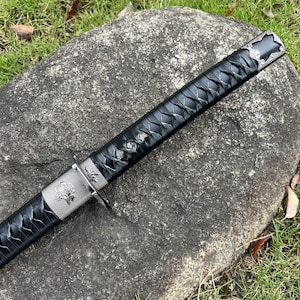 Op de afbeelding: Een zwart katana-zwaard met zilveren details. De handgreep is gewikkeld in een zwart, getextureerd materiaal. Het zwaard ligt op een grijze steen, met groen gras op de achtergrond.