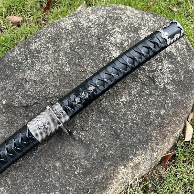 Op de afbeelding: Een zwart katana-zwaard met zilveren details. De handgreep is gewikkeld in een zwart, getextureerd materiaal. Het zwaard ligt op een grijze steen, met groen gras op de achtergrond.