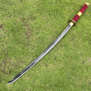 May include: A black katana sword with a red and gold handle. The sword is lying on a green grassy surface.