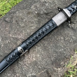 Op de afbeelding: Een zwart katana-zwaard met een zilveren bescherming en decoratieve accenten. De handgreep is gewikkeld in een textuur met een ruitpatroon. Het lemmet is niet zichtbaar. Het zwaard ligt op een grijs stenen oppervlak.