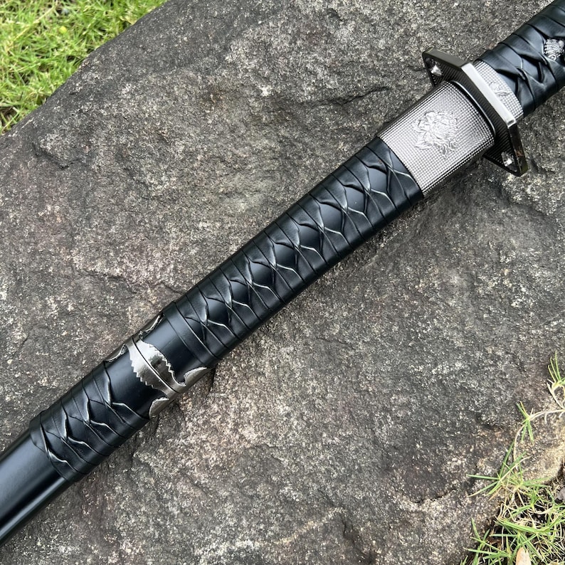 Op de afbeelding: Een zwart katana-zwaard met een zilveren bescherming en decoratieve accenten. De handgreep is gewikkeld in een textuur met een ruitpatroon. Het lemmet is niet zichtbaar. Het zwaard ligt op een grijs stenen oppervlak.