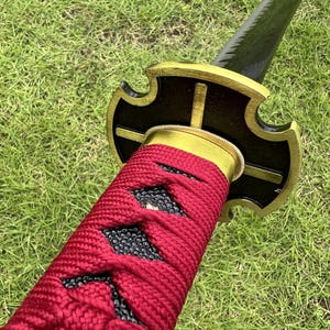 May include: Close-up of a red and black sword handle with a gold-colored metal guard. The guard has a cross-shaped design with four points.