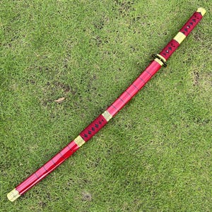 May include: A red katana with a gold-colored hilt and a black and gold patterned wrap. The katana is lying on a green grassy surface.