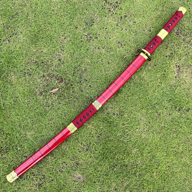 May include: A red katana with a gold-colored hilt and a black and gold patterned wrap. The katana is lying on a green grassy surface.