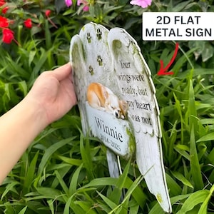 May include: A 2D flat metal sign shaped like angel wings with a printed image of a sleeping dog. The text reads: Your wings were ready, but my heart was not, Winnie, 2014-2025. The sign features small paw print graphics and a weathered stone texture.