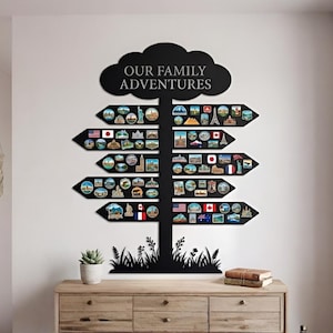 Può includere: Decorazione murale in metallo nero a forma di albero con la scritta "OUR FAMILY ADVENTURES" in alto. L'albero ha segnali direzionali con magneti a tema viaggio. Un mobile in legno e una pianta in vaso sono in primo piano.