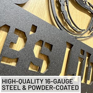 May include: A dark grey, powder-coated metal sign featuring laser-cut lettering and a circular design element. The text on the sign reads HIGH-QUALITY 16-GAUGE STEEL & POWDER-COATED. The metal surface has a textured, matte finish throughout.