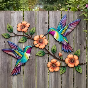 Op de afbeelding: Twee kleurrijke metalen kunstwerken van kolibries met uitgespreide vleugels, geplaatst in de buurt van oranje bloemen en groene bladeren op een houten hek. De vogels hebben levendige blauwe, groene, paarse en rode veren.