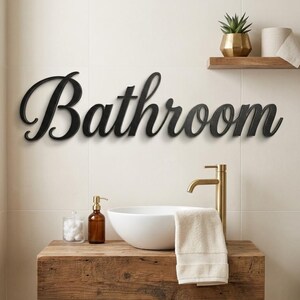 May include: A black wall-mounted sign featuring the word Bathroom in a cursive script font. The letters are connected and have a smooth finish, designed to be mounted horizontally on a flat surface to serve as a clear room identifier.