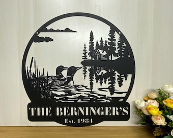 May include: Black metal wall art featuring a circular design with two loons swimming in a lake with a cabin and trees in the background. The text "The Berninger's Est. 1984" is below the image.