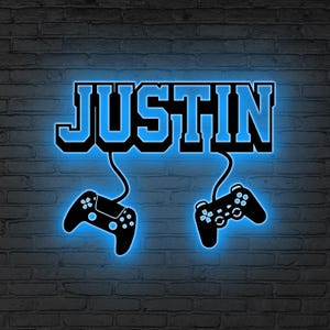 May include: A neon sign with the name "JUSTIN" in blue, outlined in black, above two black game controllers. The sign is mounted on a gray brick wall, creating a gaming theme.