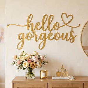 May include: A gold-coloured metal wall sign featuring the cursive text hello gorgeous with a heart symbol at the end. The sign is mounted on a plain wall above a wooden surface holding a glass vase with a bouquet of white, pink, and tan flowers.