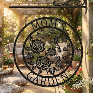 以下が含まれることがあります： 「MOM'S GARDEN」の文字が入った黒い金属製のガーデンサイン。円形のデザインで、バラ、花、ハチドリ、葉の切り抜き画像が特徴です。装飾的なブラケットから吊り下げられています。