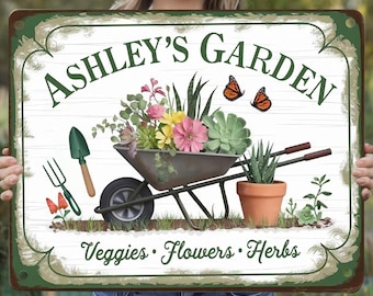 Könnte beinhalten: Ein grünes und weißes Metallschild mit dem Text "Ashley's Garden" in einer dekorativen Schriftart. Das Schild zeigt eine Schubkarre gefüllt mit Blumen, Pflanzen und Gartenwerkzeugen. Der Text "Veggies, Flowers, Herbs" befindet sich am unteren Rand des Schildes.