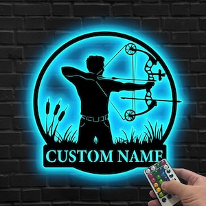 Peut inclure: Décoration murale en métal noir représentant un archer tirant à l'arc, entouré d'un cercle bleu lumineux. Le design comprend de l'herbe et des roseaux, avec le texte "CUSTOM NAME" en dessous. Une télécommande est visible.