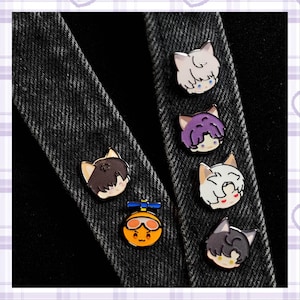 May include: A selection of enamel pins showcasing cartoon cat faces with various hairstyles and colours. One pin features a smiling orange character with goggles. The pins are displayed on a dark denim background.
