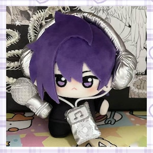 Puede incluir: Una muñeca de peluche con cabello morado, que lleva auriculares plateados y un atuendo negro. La muñeca sostiene un micrófono plateado y un reproductor de música plateado con un símbolo de nota musical. La muñeca tiene ojos grandes y expresivos y una linda expresión.