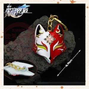 May include: A red, white, and gold fox mask keychain. The mask is split vertically, with a red side and a white side. The keychain measures 4.5 cm in length. The mask has gold accents and a gold chain.