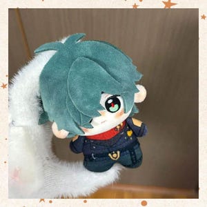 May include: A plush doll with teal hair and large, expressive eyes. The doll wears a dark blue outfit with gold accents and a red scarf. The doll is attached to a white, fluffy hair accessory.