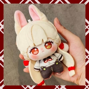 May include: A plush doll with a bunny-like appearance, featuring cream-colored fur, pink inner ears, and long, flowing hair. The doll has a detailed face with large, expressive eyes and is wearing a white and black outfit with brown pants. Red accents are present.