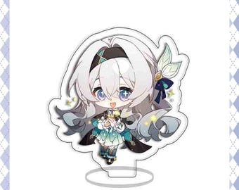 May include: A cartoon acrylic stand featuring a chibi anime character with white hair, blue eyes, and a teal and black outfit. The character is holding a small object and has a cheerful expression. The stand has a clear base.