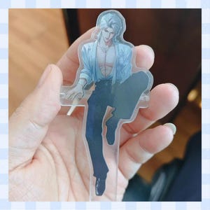 May include: A clear acrylic standee depicting a male anime character with silver hair, wearing a light blue open shirt and dark blue trousers. The character is in a seated pose. The standee is held in a hand.