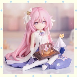May include: A detailed figurine of a seated anime-style character with long pink hair and purple eyes. The figure wears a white and blue outfit and holds a miniature book with a stage set. A small bird figure sits atop a white cloth.