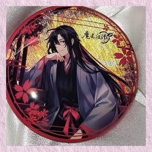 May include: A round, red and gold decorative item featuring an anime-style illustration of a person with long black hair and a dark outfit. The item has a red border with floral accents and text in the upper right corner.