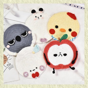 Peut inclure: Une collection de cinq dessous de verre crochetés à la main, de formes et de couleurs variées. Les dessous de verre comprennent un koala gris, un lapin blanc, un poussin jaune, une pomme rouge et un personnage blanc avec un nœud rouge. Chaque dessous de verre présente des détails brodés.