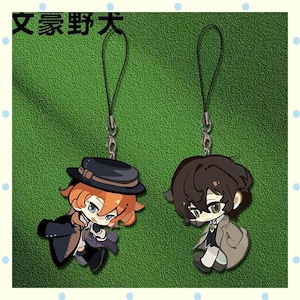 Peut inclure: Deux breloques de personnages de style anime avec des lanières noires. L'une représente un personnage aux cheveux orange et un chapeau noir, l'autre a les cheveux bruns. Le fond est vert avec le texte japonais "Bungo Stray Dogs" en haut.