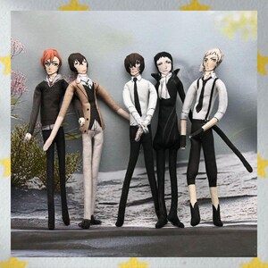 May include: Five handmade fabric dolls, each dressed in unique outfits. The dolls have detailed facial features and hairstyles. The outfits include suits, shirts, and ties in various colors. The dolls are standing in a row against a blurred background.