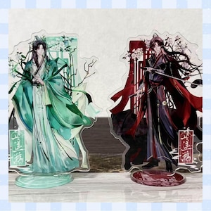 May include: Two acrylic anime character standees. One depicts a figure in green robes, the other in red and black robes, holding a sword. Both have decorative bases and floral accents. The figures have long black hair.
