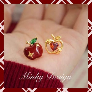 Puede incluir: Dos pendientes con forma de manzana. Uno es rojo brillante con detalles de estrellas doradas y una hoja verde. El otro es dorado, con un centro de gema roja en forma de corazón. Los pendientes se muestran en una mano, con el texto "Minky Design" en la parte inferior.
