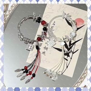 Könnte beinhalten: Zwei verzierte silberne Armbänder mit Schmetterlingsakzenten, roten und schwarzen Perlen und Quasten. Ein Armband hat einen quadratischen Anhänger mit chinesischen Schriftzeichen. Die Armbänder werden auf einer reflektierenden Oberfläche präsentiert.