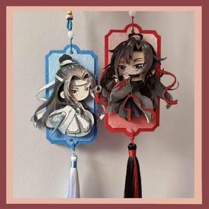 May include: Two anime-style paper ornaments. One is blue with a white and grey character, the other is red with a black and red character. Both have tassels and are hanging.