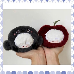 Puede incluir: Dos peluches: una foca gris oscuro con cara blanca y una cereza roja. Ambos tienen caras bordadas. Los juguetes se sostienen en las manos.