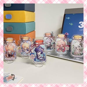 Puede incluir: Soportes acrílicos con forma de tarro, con ilustraciones de estilo anime. Cada soporte presenta un diseño de personaje único con colores vibrantes y pequeñas etiquetas. Los tarros tienen tapas de corcho y están atados con cintas rojas.