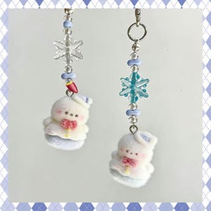 May include: Two winter-themed keychains. Each keychain features a fluffy white bunny with a red bow, a blue hat, and a snowflake charm. The keychains are adorned with blue and clear beads and a silver clasp.